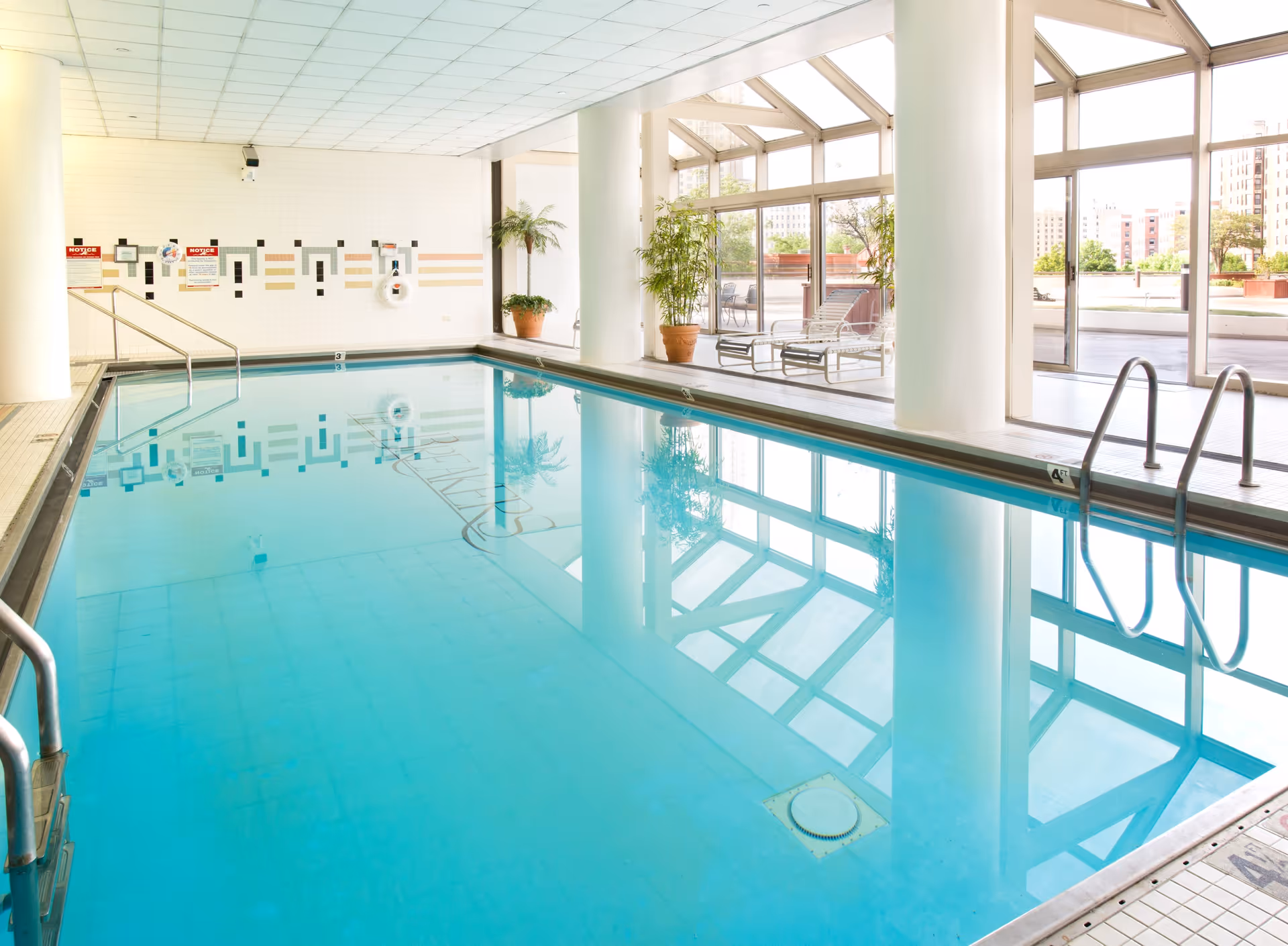 Indoor swimming pool with clear blue water, surrounded by white tiled walls and large windows letting in natural light. There are potted plants and lounge chairs near the windows, with a view of an outdoor patio area and city buildings in the background.