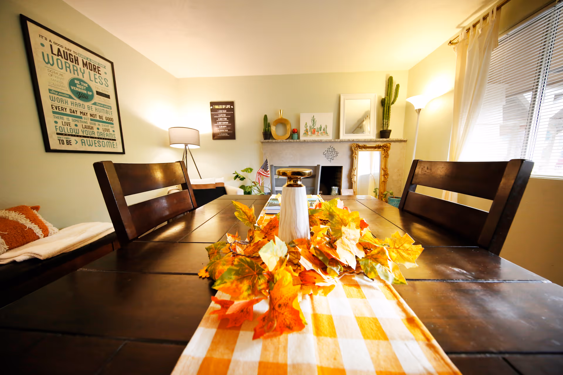 A cozy dining room with a dark wooden table decorated with a yellow and white checkered runner and autumn leaves centerpiece. The room has wooden chairs, a lamp in the corner, framed inspirational quotes and artwork on the walls, a fireplace with decorative items, a tall cactus plant, and a window with blinds and curtains letting in natural light.