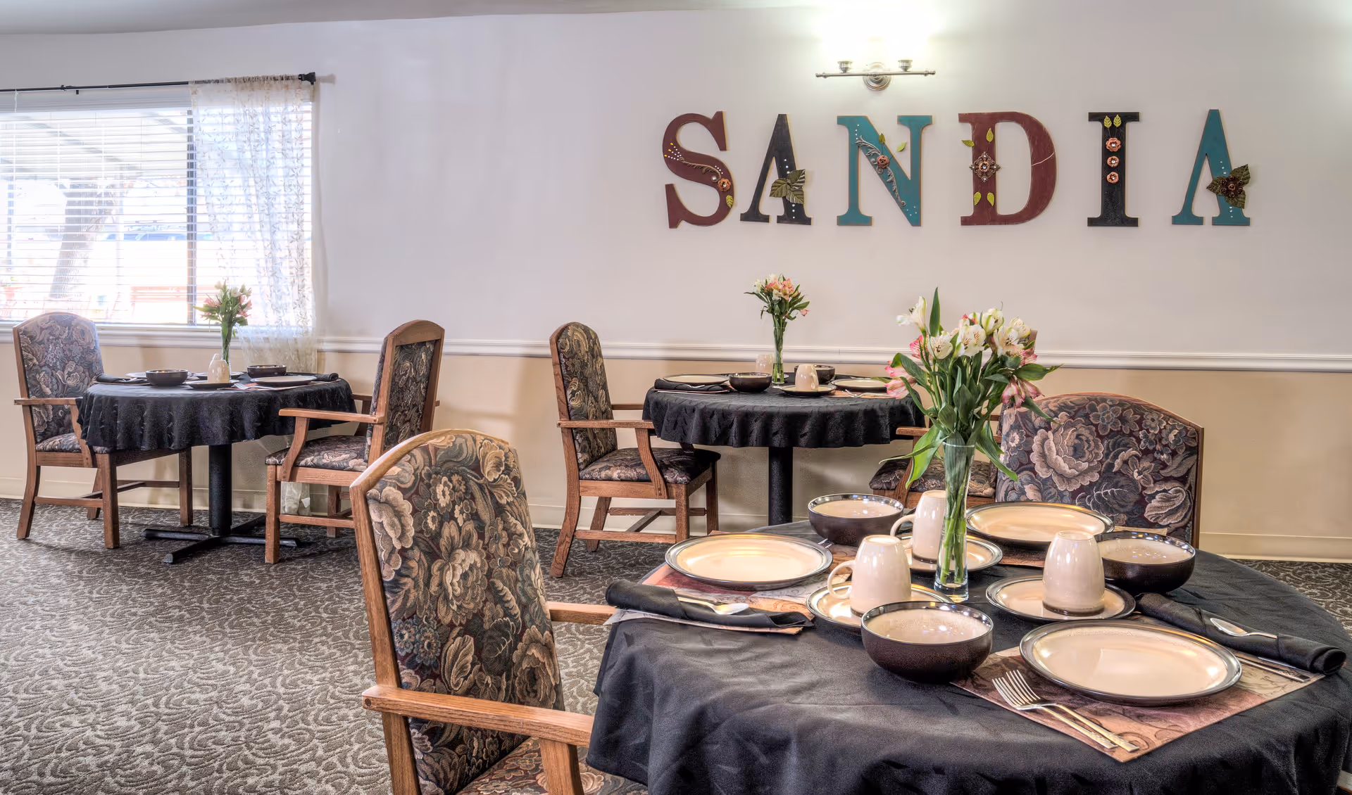 Dining room with round tables covered in black tablecloths, each set with plates, bowls, cups, and silverware. Floral arrangements in vases are placed on each table. Upholstered chairs with floral patterns surround the tables. The wall has decorative letters spelling 'SANDIA'. A window with blinds and sheer curtains lets in natural light.