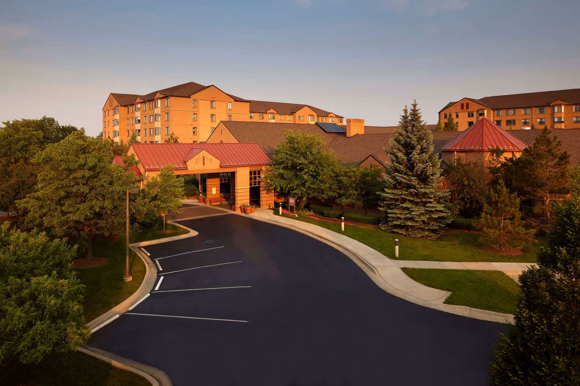 Exterior view of Allegria Village senior living facility during sunset, showing a large brick building with multiple floors, a driveway with parking spaces, and surrounding trees and greenery.