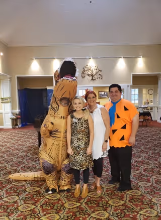 Four people standing indoors on a patterned carpet. One person is wearing an inflatable dinosaur costume, another is dressed in a leopard print outfit, the third person is wearing a white sleeveless top with black pants, and the fourth person is dressed as Fred Flintstone in an orange tunic with black triangles and a blue tie. The background shows a beige wall with decorative lighting and a tree wall art.