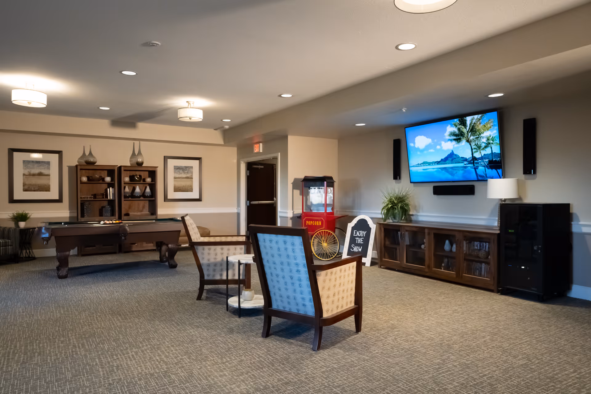 A spacious common area with two upholstered chairs facing a large wall-mounted TV displaying a tropical beach scene. There is a popcorn machine, a sign that says 'Enjoy the Show,' a pool table, and wooden shelving with decorative items. The room has soft lighting and neutral-colored walls and carpet.