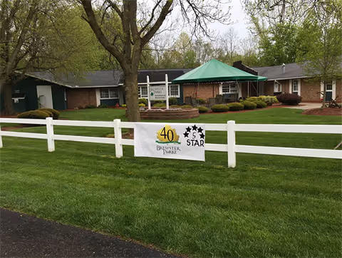 Exterior view of Brewster Parke, Inc facility showing a single-story brick building with a green canopy over the entrance. There is a white fence in the foreground with a banner celebrating 40 years and a 5-star rating. The lawn is well-maintained with trees and shrubs around the building.