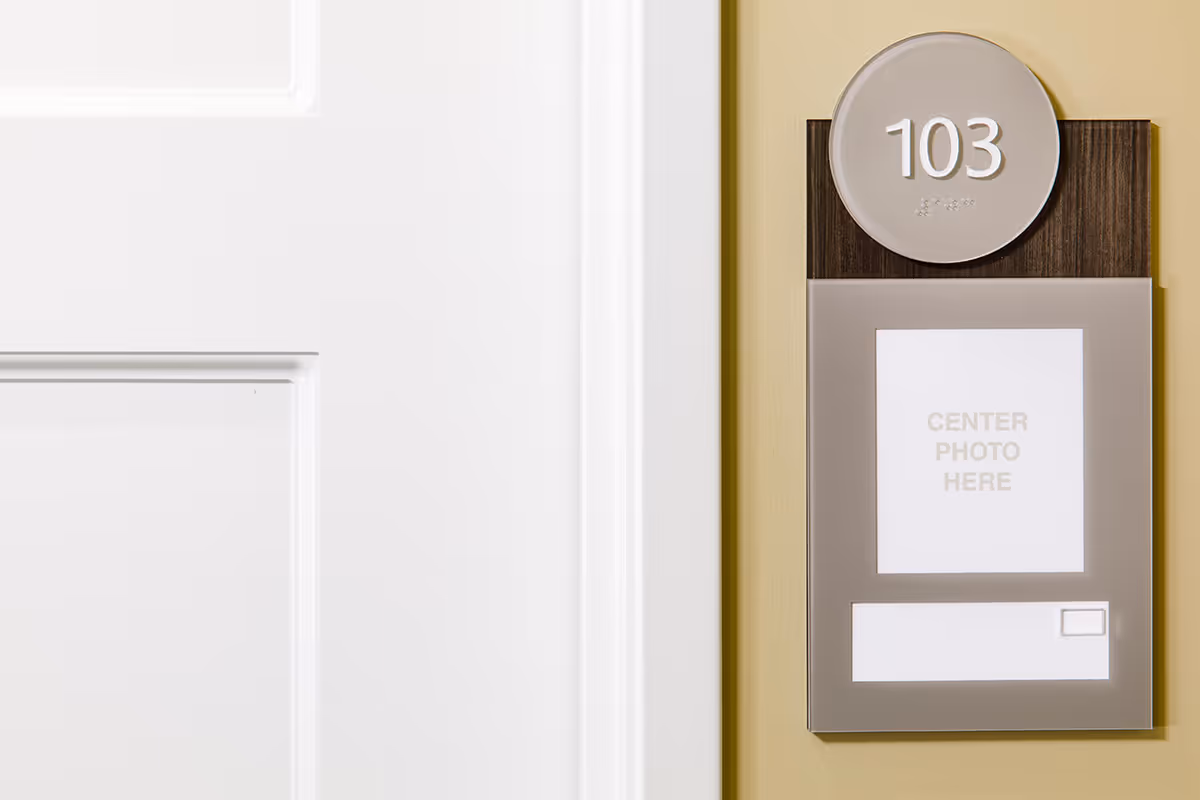 A close-up view of a room number sign mounted on a beige wall next to a white door. The sign displays the number 103 in large white text on a circular taupe background with Braille below it. Below the circle is a rectangular section with a placeholder text that reads 'CENTER PHOTO HERE'.