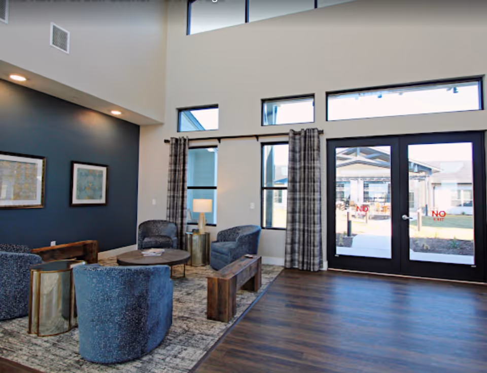 Bright communal lounge with blue upholstered chairs around a round coffee table, tall windows and glass double doors leading outside.