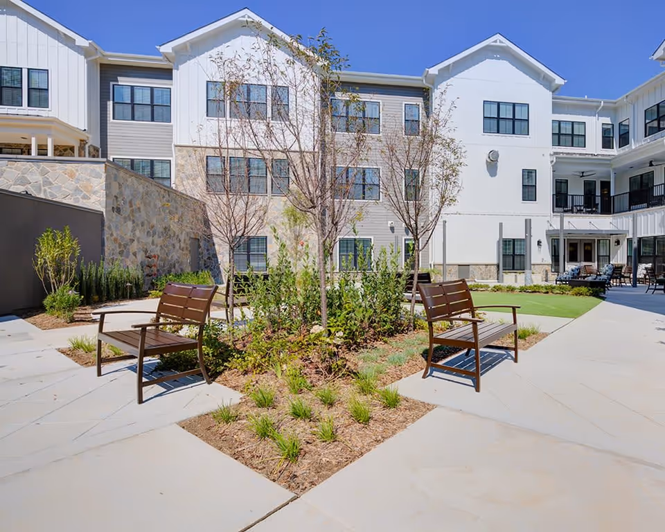 Outdoor courtyard area at The Residence at Colvin Run featuring two wooden benches facing each other around a landscaped garden bed with small trees and plants. The courtyard is surrounded by a multi-story building with white and stone exterior walls, multiple windows, and a clear blue sky overhead.