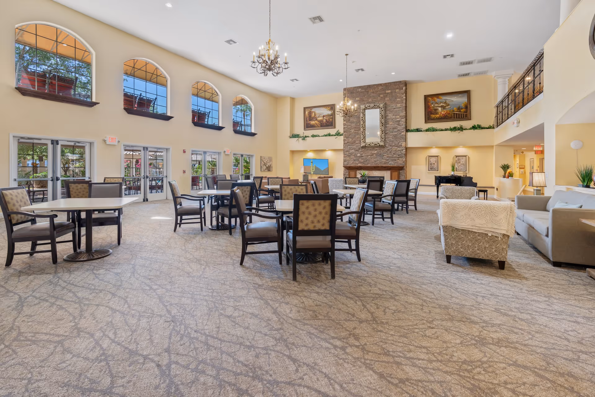 Spacious senior living common area with multiple tables and chairs arranged for socializing or dining, large windows with arched tops letting in natural light, a stone fireplace with a mirror above it, chandeliers hanging from the ceiling, and comfortable seating including sofas and armchairs.