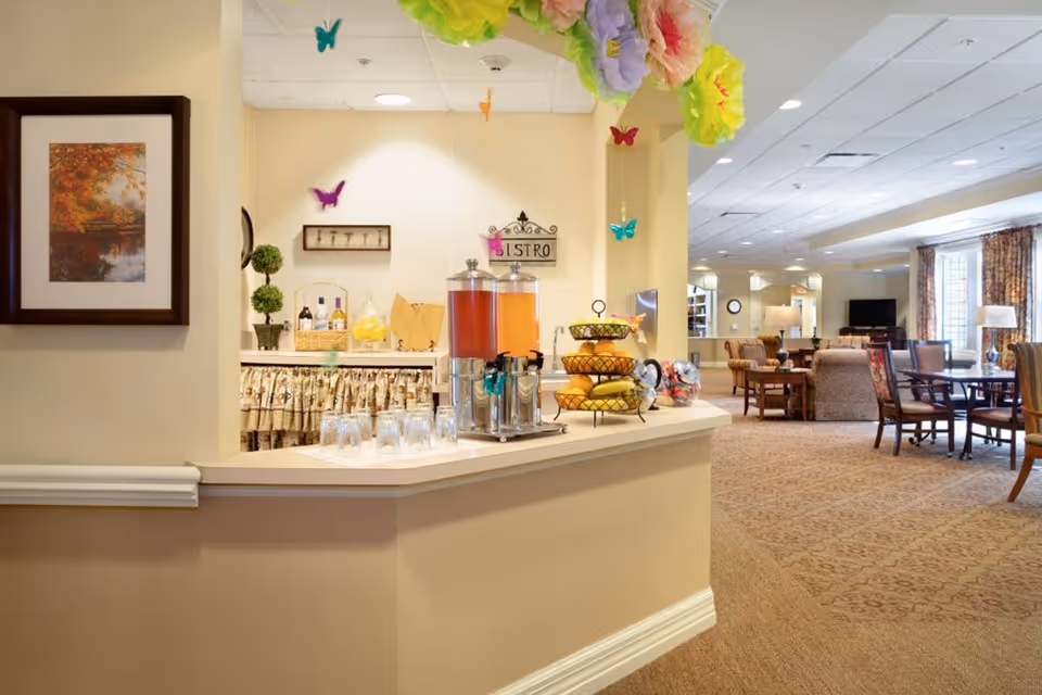 Interior view of a senior living facility featuring a small refreshment area with beverage dispensers, glasses, and a tiered fruit basket on a counter. The background shows a spacious common area with tables, chairs, armchairs, a television, and large windows with curtains. Decorative paper flowers and butterflies hang from the ceiling near the refreshment area.