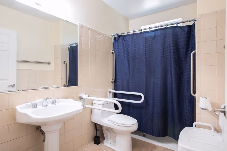 A clean bathroom with beige tiled walls and floor, featuring a white pedestal sink with chrome faucets, a toilet with white safety grab bars on both sides, a dark blue shower curtain, and a white shower chair inside the shower area. There is a large mirror above the sink and a towel bar on the wall.
