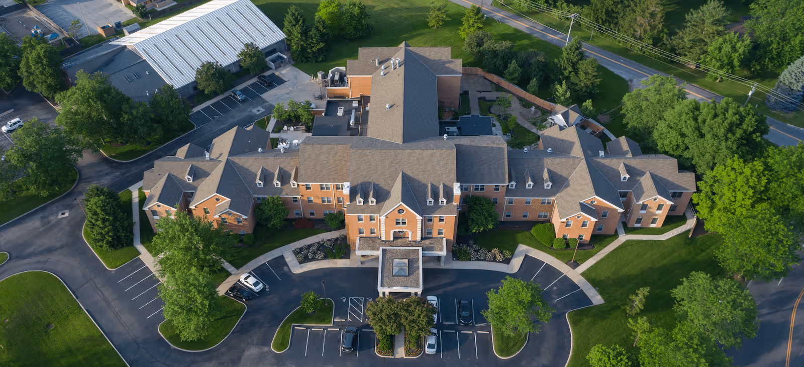 Aerial view of a large senior living facility building with multiple wings, surrounded by green lawns, trees, and parking areas with several cars parked. The building has a brick exterior and a gray roof, with pathways and landscaped areas around it.