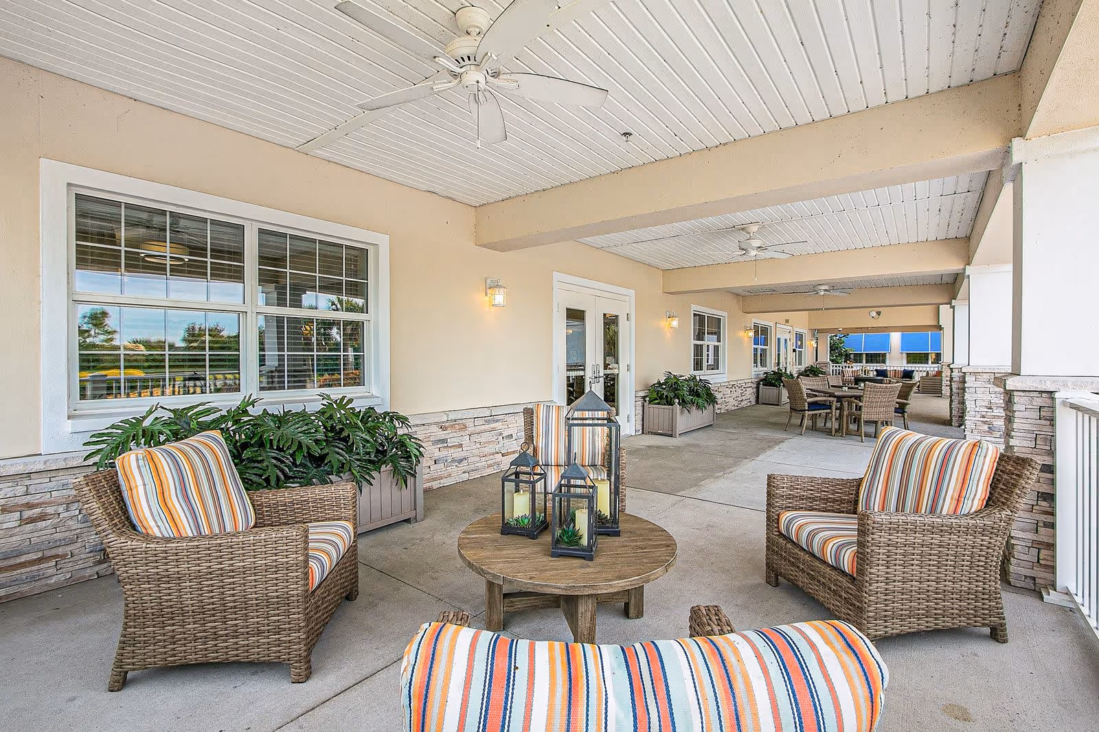 Covered outdoor patio area with wicker chairs featuring striped cushions arranged around a wooden round table with decorative lanterns. The patio has ceiling fans, potted plants, and multiple seating areas with tables and chairs along the side of a building with windows and doors.