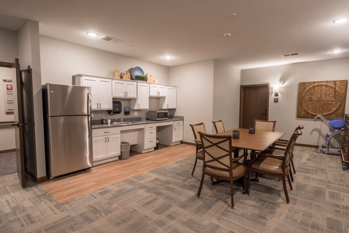Communal dining area with a kitchenette featuring a stainless steel refrigerator, white cabinets, and a table with six chairs.