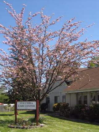 Single-story care center building with a blooming pink-flowered tree and a lawn sign that reads "Columbia Care Center".