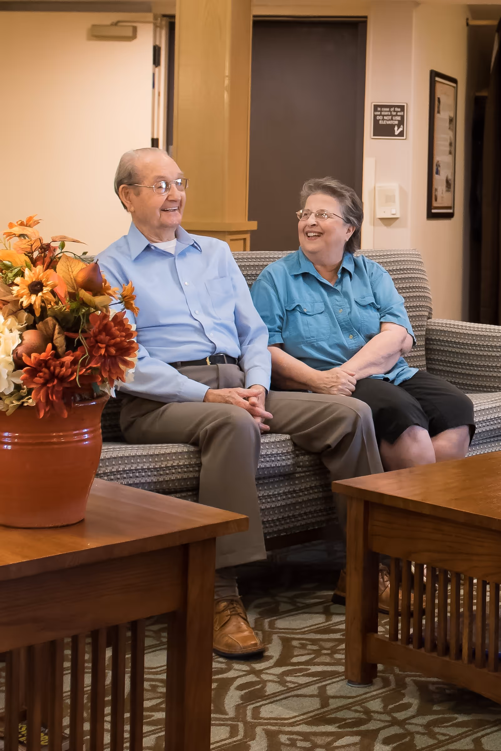 An elderly man and woman sitting on a patterned couch in a cozy living room, smiling and looking at each other. There is a wooden coffee table with a large vase of colorful flowers in the foreground, and a patterned carpet on the floor.