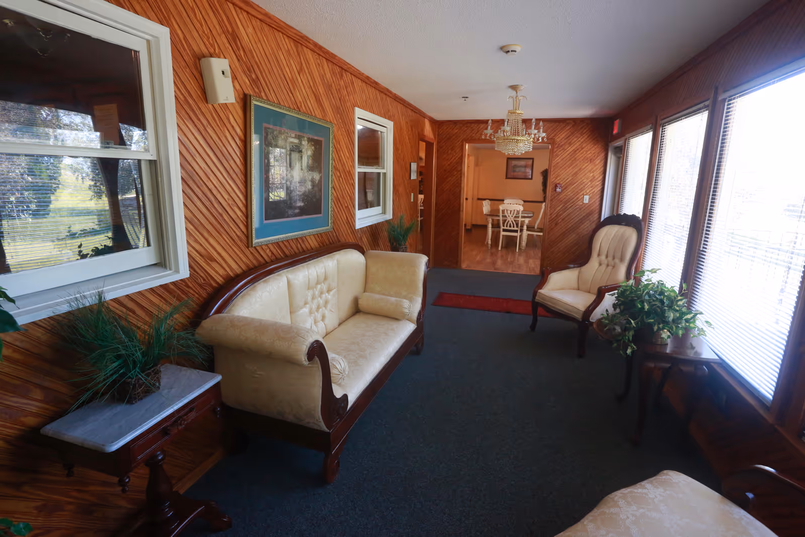 A cozy sitting area with vintage-style cream upholstered sofa and armchair, wooden paneled walls, a small side table with a plant, and large windows with blinds letting in natural light. A chandelier hangs from the ceiling, and a dining area with a table and chairs is visible through an open doorway.