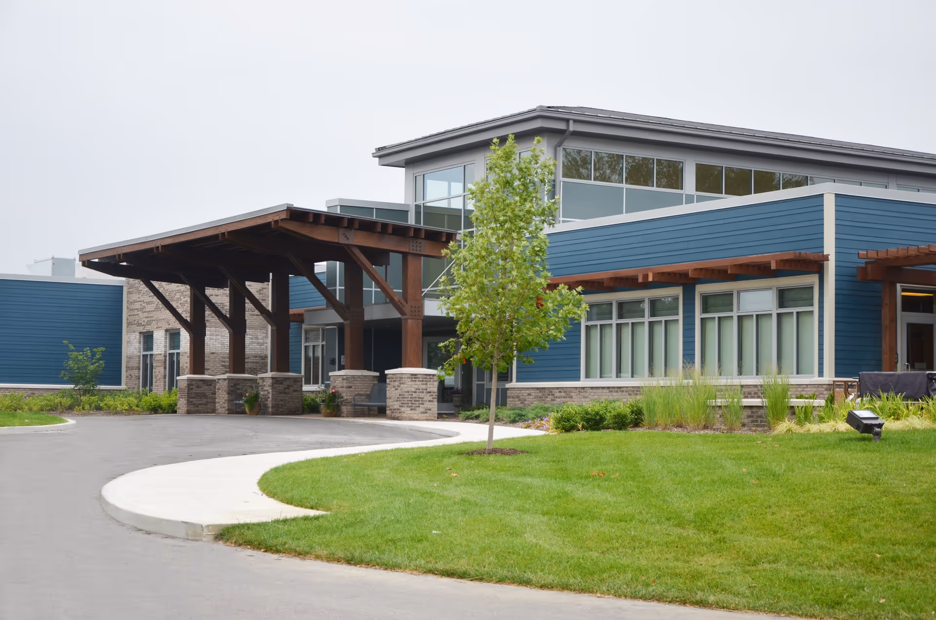 Exterior view of Arlington Place Health Campus showing a modern building with blue siding and large windows. There is a covered entrance with wooden beams and brick pillars, a curved driveway, green lawn, and a young tree in front.