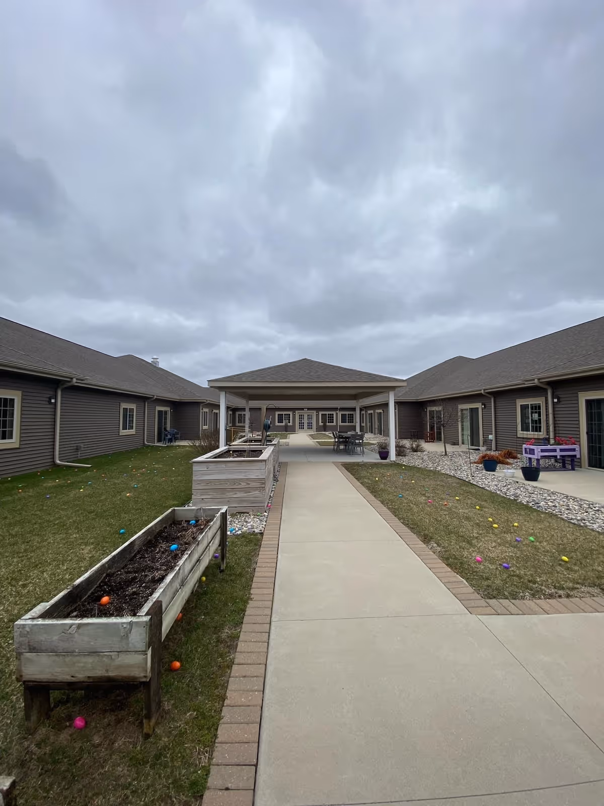 Outdoor courtyard area of a senior living facility with a paved walkway leading to a covered seating area. Raised garden beds are on the left side of the walkway, and colorful plastic Easter eggs are scattered on the grass on both sides. The building has brown siding and multiple windows and doors facing the courtyard under a cloudy sky.