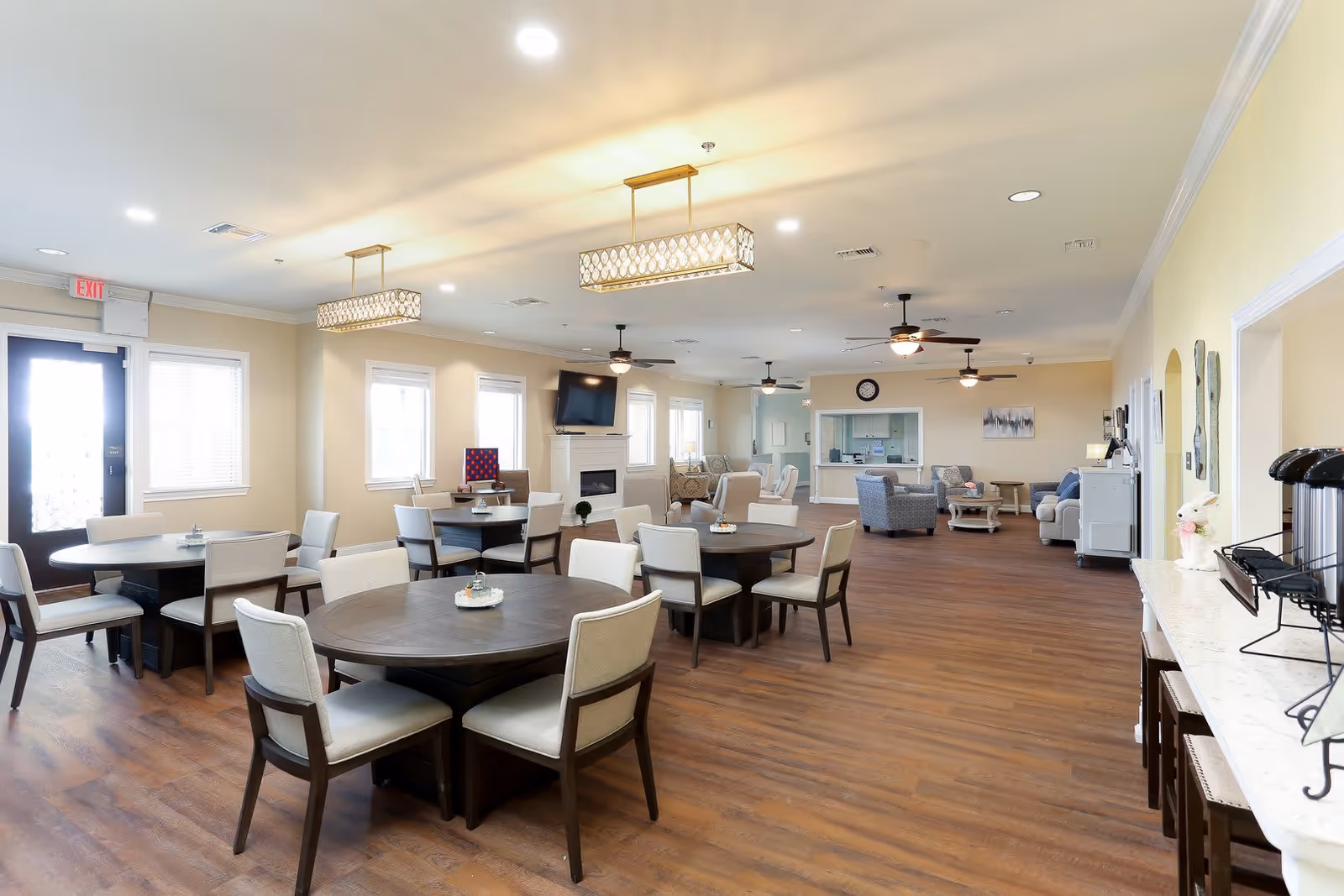 A spacious and well-lit common area in an assisted living facility featuring several round wooden tables with cushioned chairs arranged around them. The room has a warm wooden floor, beige walls, and multiple windows allowing natural light. There are modern ceiling lights and ceiling fans, a wall-mounted TV above a white fireplace, and a seating area with armchairs and sofas in the background. A counter with coffee dispensers and stools is visible on the right side.