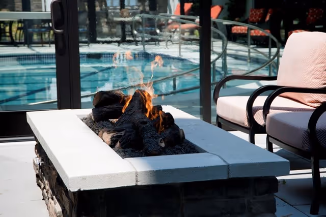 A modern outdoor fire pit with burning logs surrounded by cushioned patio chairs, with a swimming pool and glass doors in the background.
