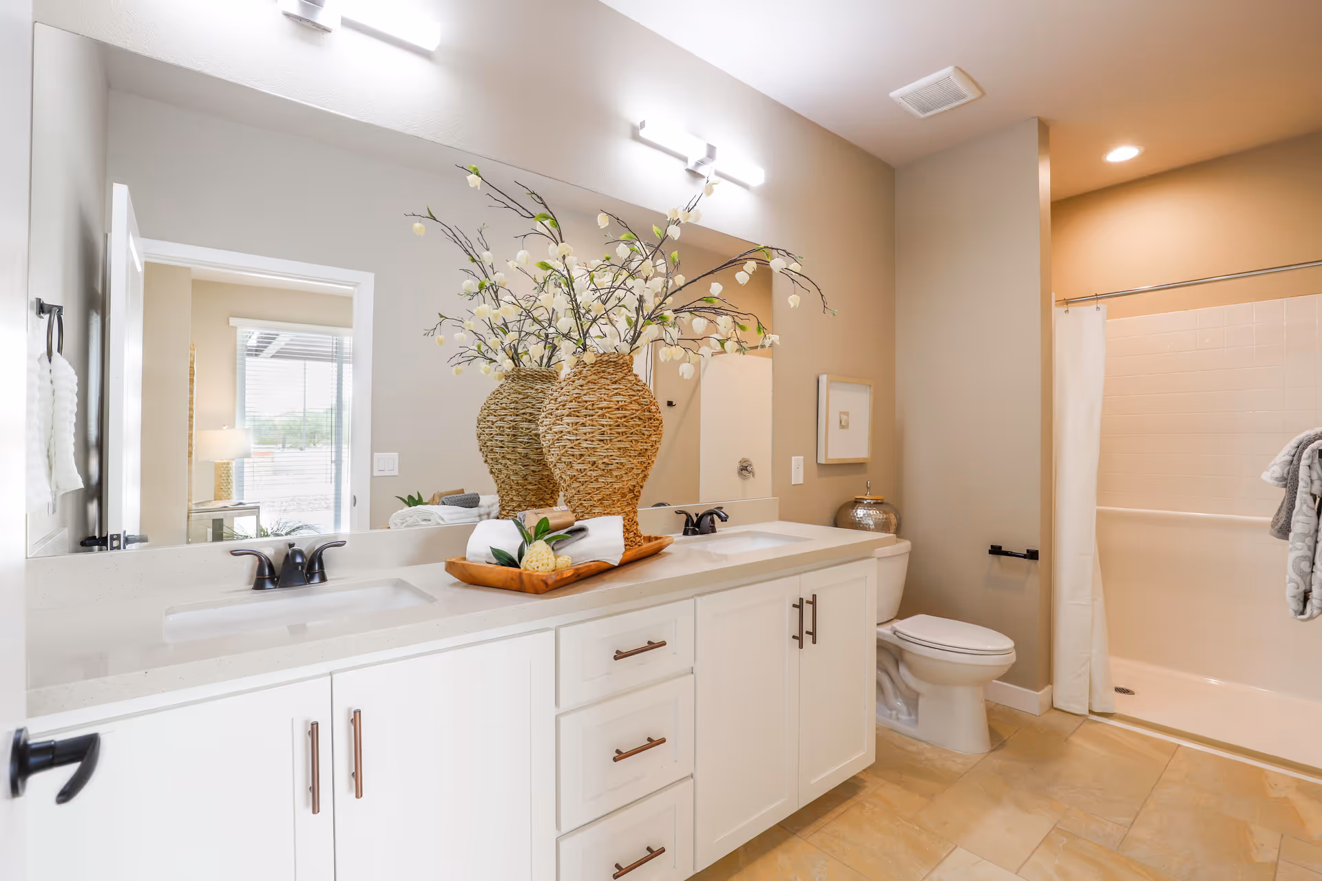 Bright and clean bathroom with a double sink vanity featuring white cabinets and a large mirror. A decorative vase with flowers sits on the countertop along with neatly rolled towels. The bathroom includes a toilet and a shower with a white curtain. The walls are painted beige and the floor is tiled.