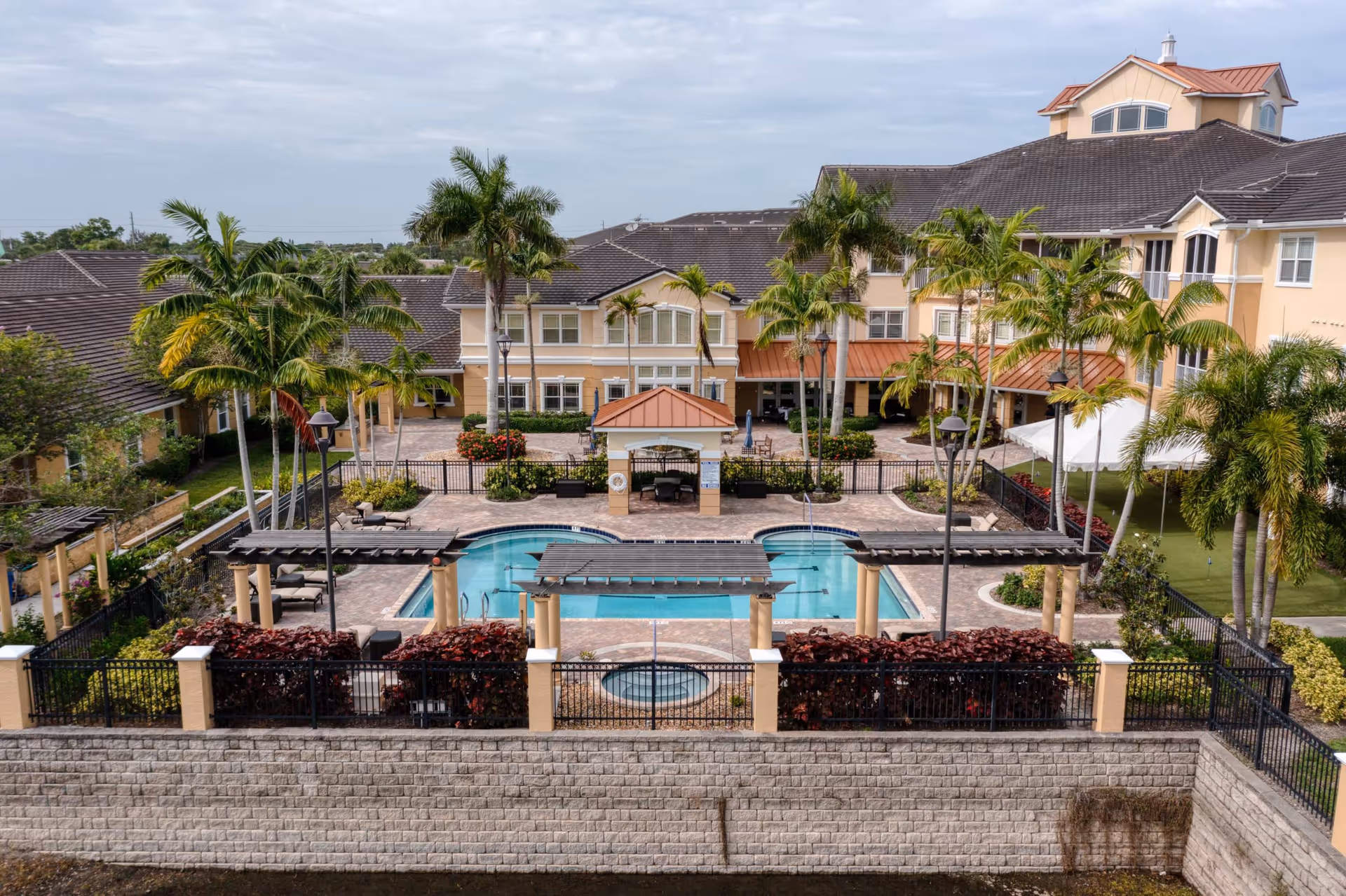 Outdoor view of Addington Place of Stuart featuring a swimming pool surrounded by palm trees, lounge chairs, and a pergola. The building with multiple windows and a tiled roof is visible in the background under a cloudy sky.