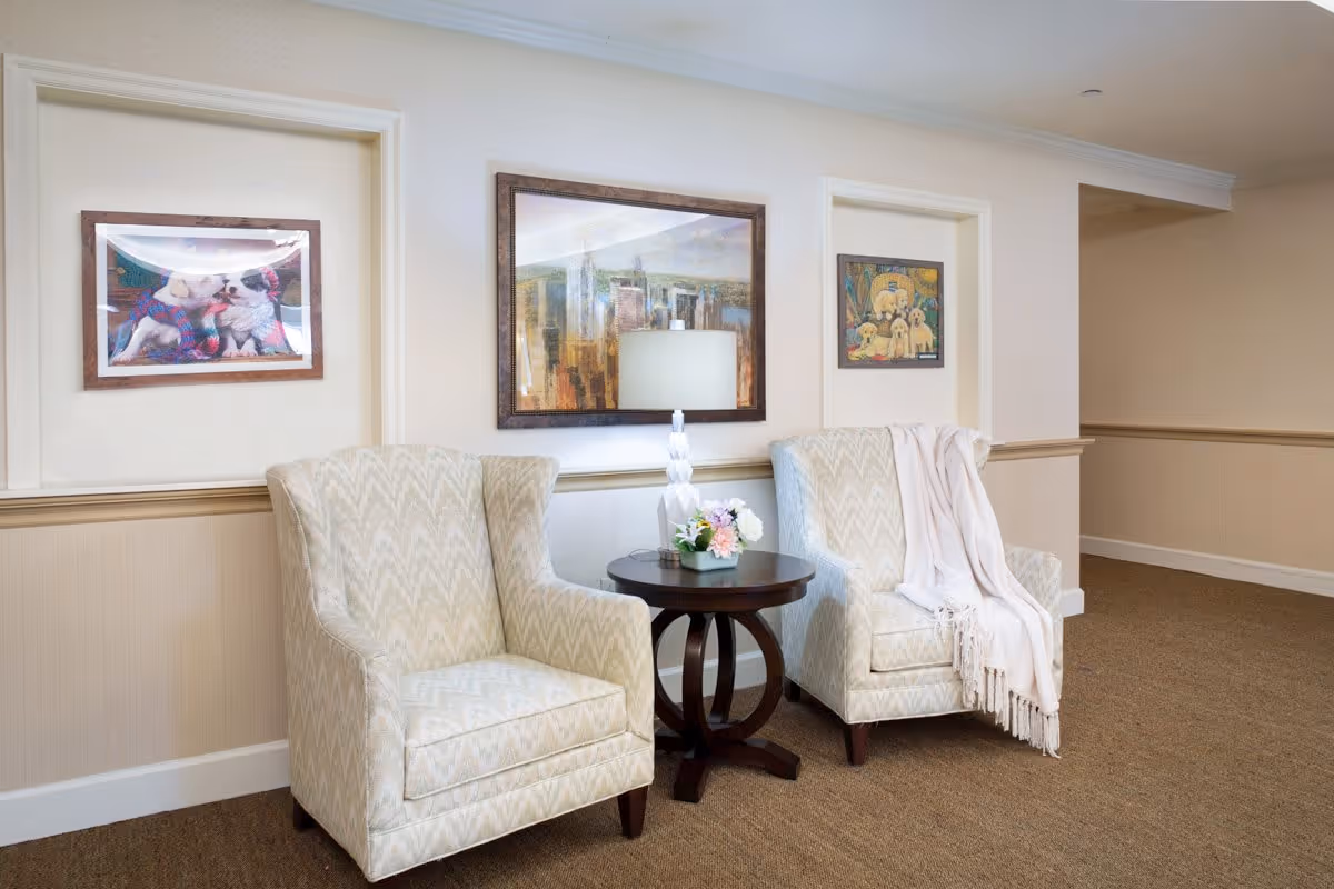 Two patterned armchairs with a white throw blanket on one chair, positioned on either side of a small round wooden table with a white lamp and a small flower arrangement. The wall behind features three framed pictures, including images of dogs and a cityscape.