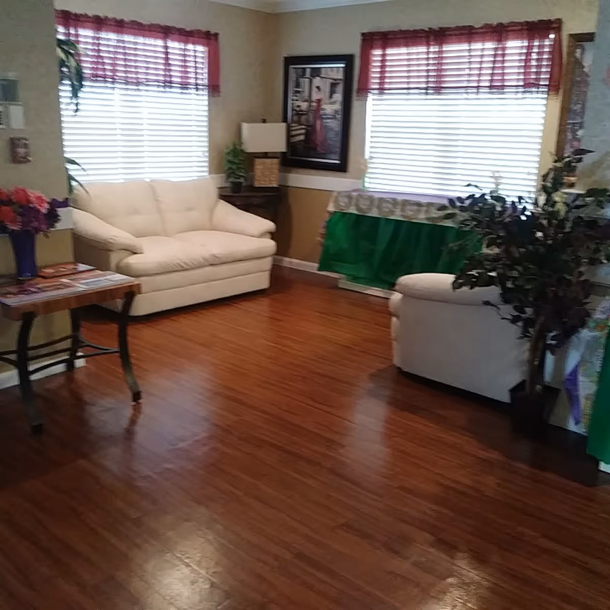 A cozy living room with two white sofas, wooden flooring, two windows with red valances and blinds, a small wooden table with a vase of flowers, a framed picture on the wall, a lamp on a side table, and some green plants.