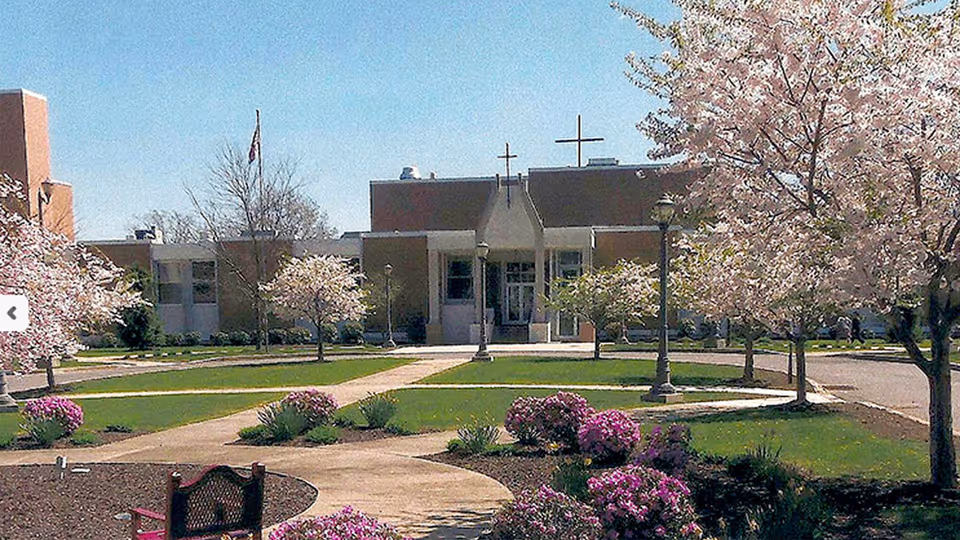 Front exterior of a care facility with a lawn, walkways, flowering trees, lamp posts, and a central entrance topped by crosses.