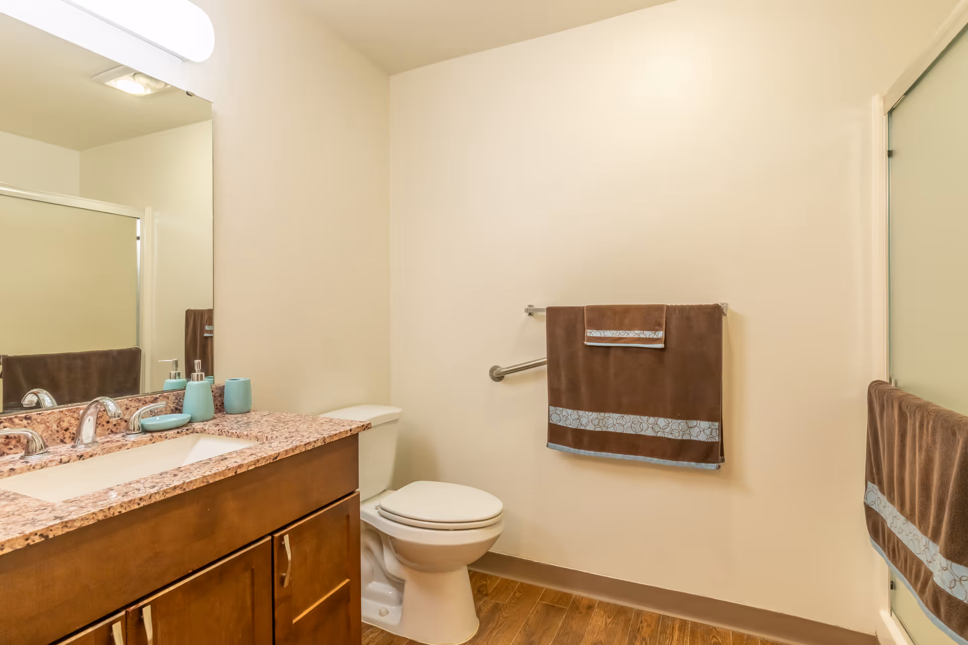 A clean bathroom with a wooden vanity topped with a speckled granite countertop, a sink with chrome faucet, a large mirror, a white toilet, and brown towels with light blue decorative trim hanging on the wall and shower door. The floor has wood-like tiles and the walls are painted light beige.