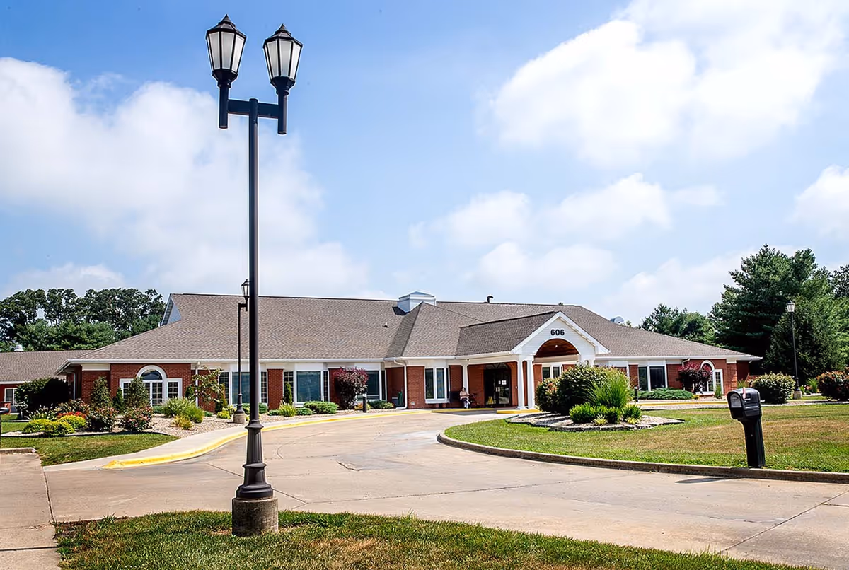 Exterior view of Mt Vernon Countryside Manor, a single-story brick building with a peaked roof, surrounded by landscaped greenery and a curved driveway. A black lamppost with two lanterns stands in the foreground under a partly cloudy sky.