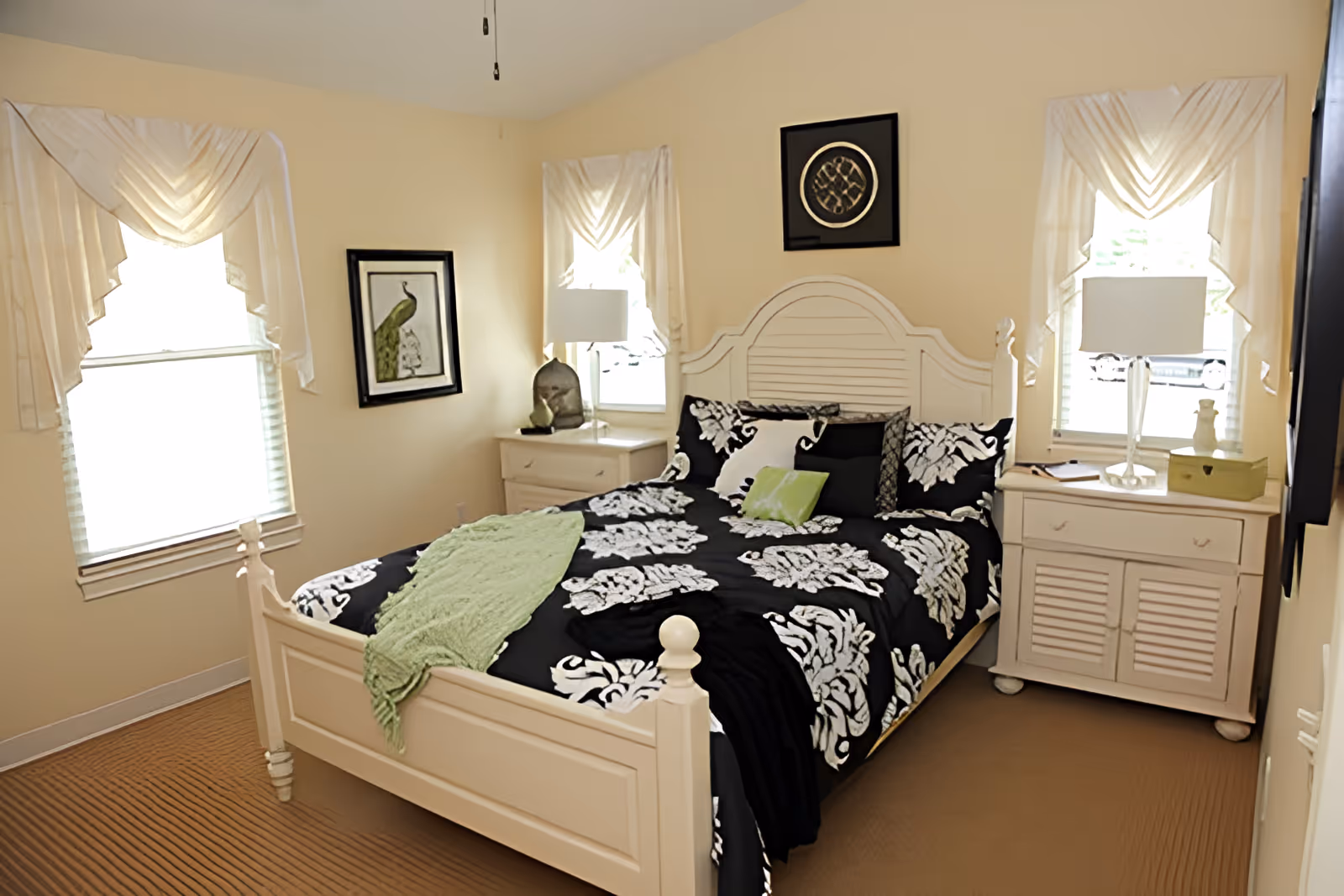 A bedroom with a white wooden bed frame and a black and white patterned bedspread with green accent pillows and a green throw. The room has beige walls, two windows with white curtains, two white nightstands with lamps, and framed artwork on the walls.