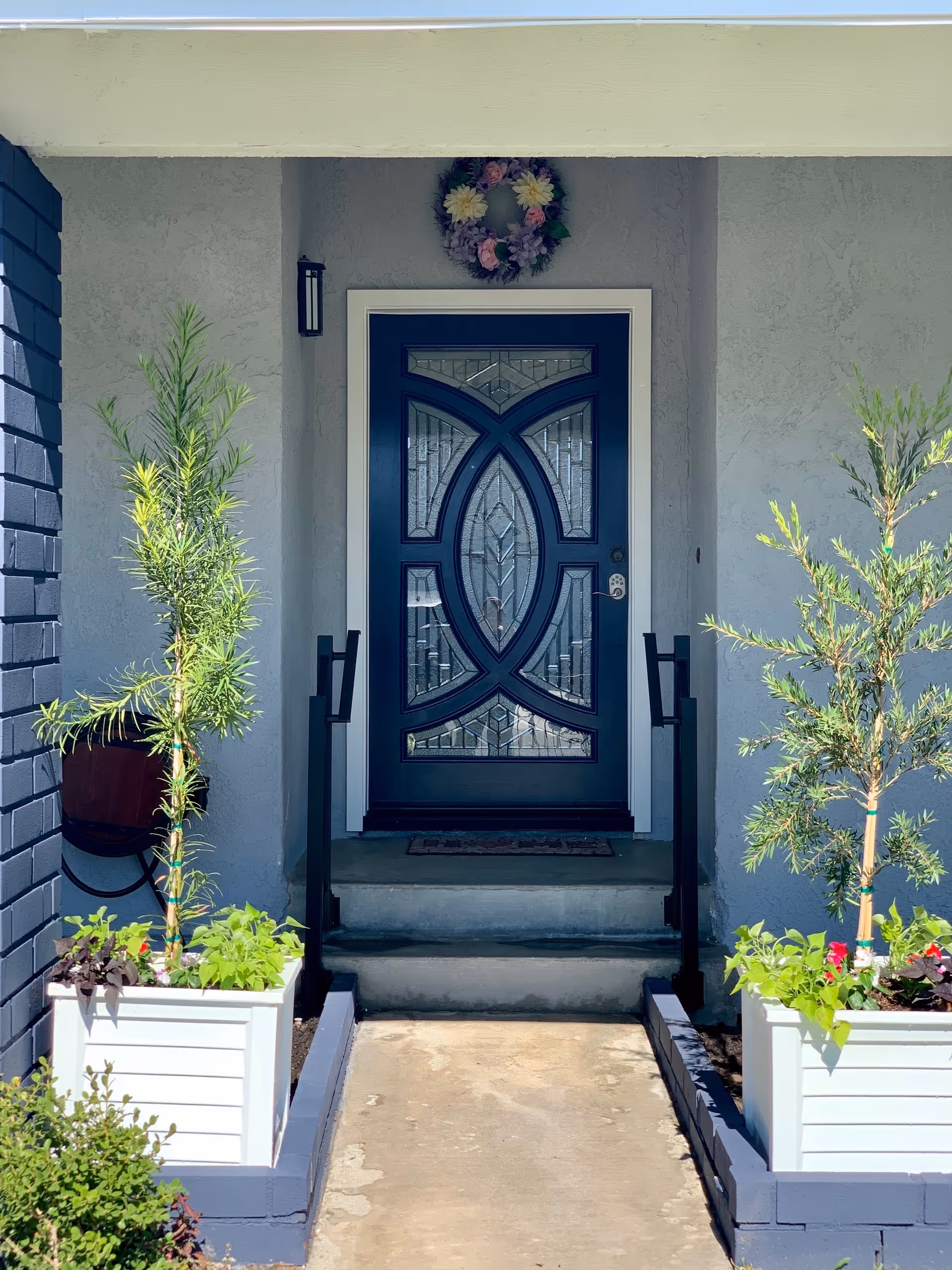 Front entrance of a building with a decorative dark blue door featuring intricate glass panels. The entrance has two steps leading up to the door, black handrails on both sides, and two white planter boxes with green plants and small trees on either side of the walkway. A floral wreath hangs above the door on a light gray textured wall.