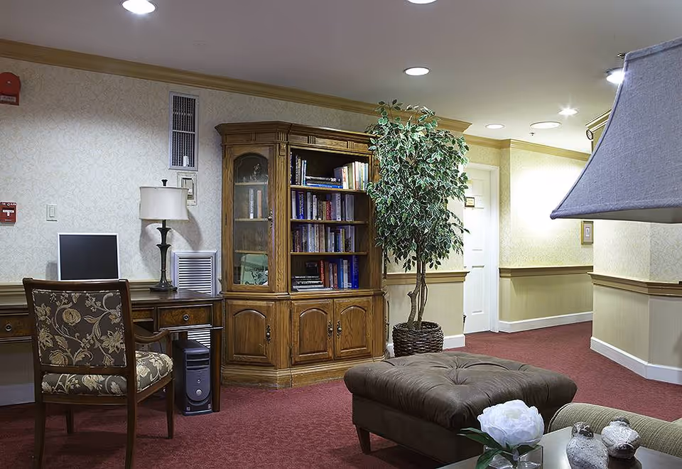 Interior of a senior living facility room with a wooden bookshelf filled with books, a desk with a computer and lamp, a patterned chair, a large potted plant, a brown cushioned ottoman, and a table with a white flower and decorative items. The room has beige wallpaper, red carpet, and recessed ceiling lights.