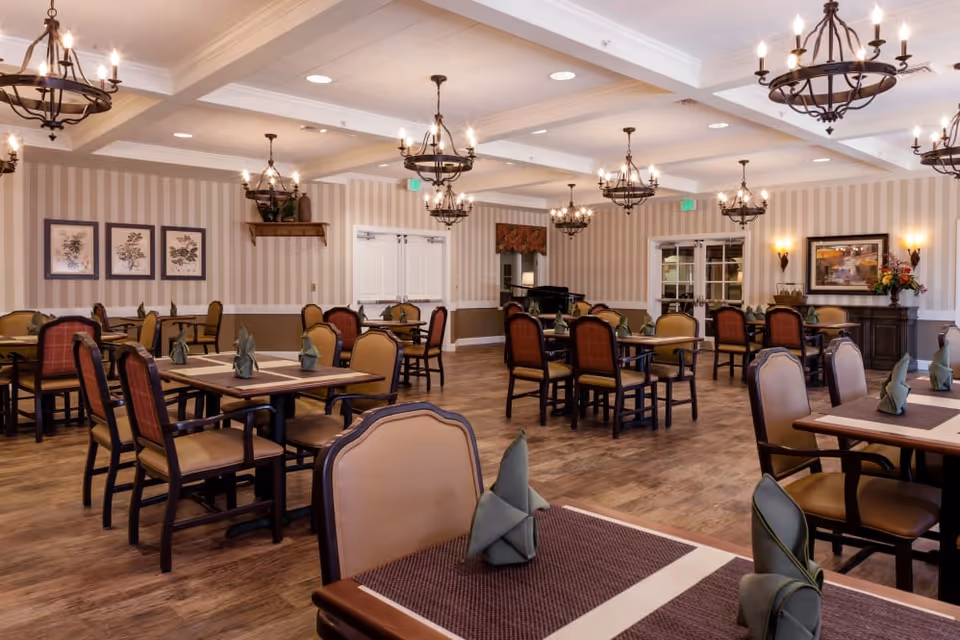 A spacious dining room with multiple tables and chairs arranged neatly. Each table has a placemat and a folded green napkin. The room features wooden flooring, striped wallpaper, framed botanical artwork on the walls, and several elegant chandeliers hanging from the ceiling. There is a sideboard with a flower arrangement and a framed picture, and double doors leading to another area.