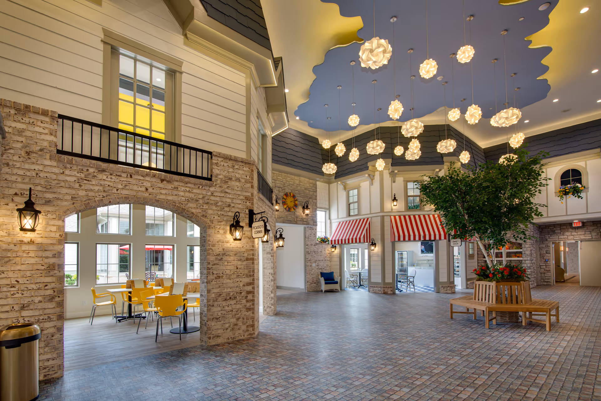 Spacious indoor common area with brick archways, a seating area of yellow chairs, a central planter bench with a tree, and decorative hanging pendant lights under a sculpted blue ceiling.