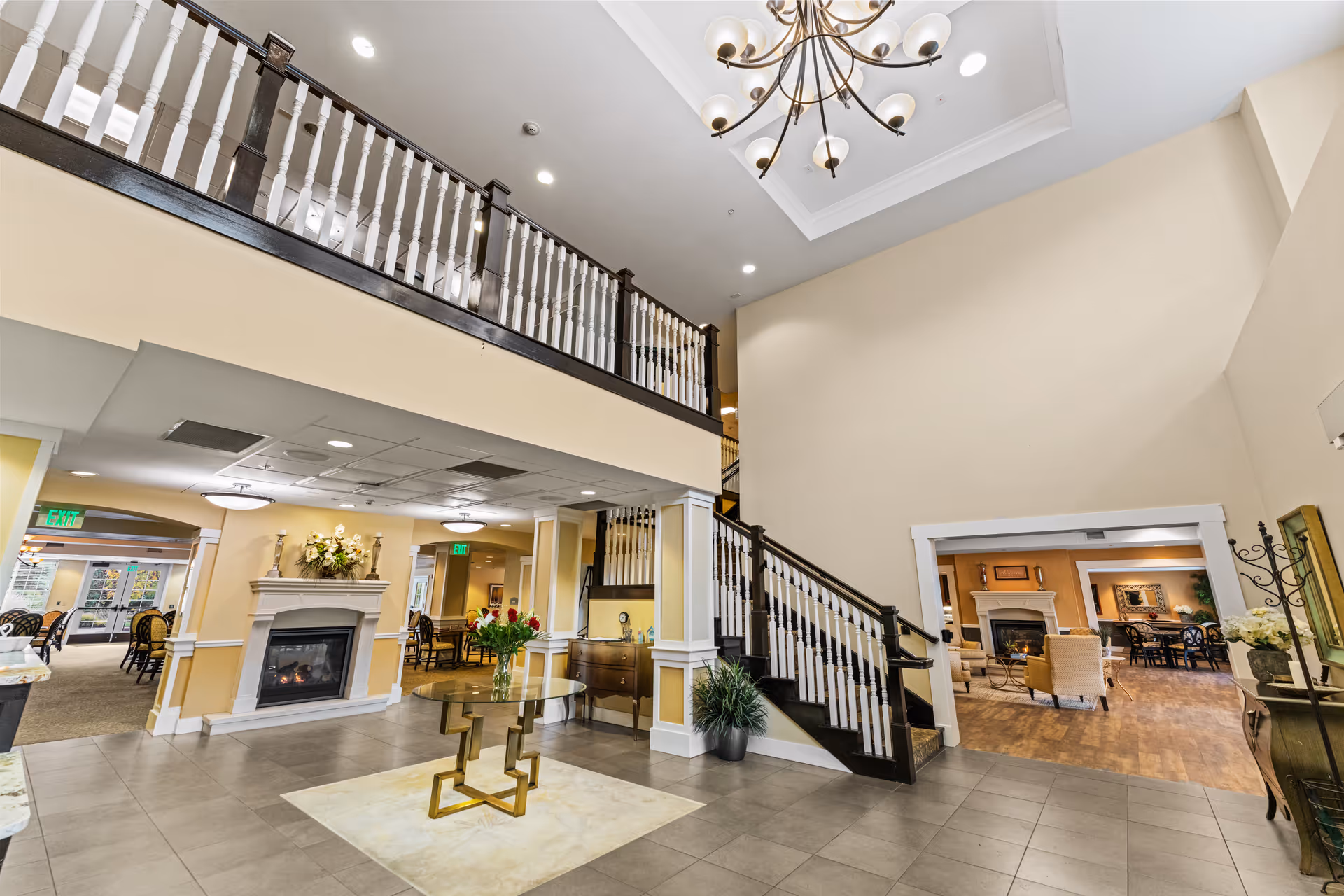 Spacious and well-lit interior of a senior living facility featuring a high ceiling with a chandelier, a staircase with dark wooden railings and white balusters, a glass table with a flower vase in the center, a fireplace in the background, and seating areas visible through doorways.
