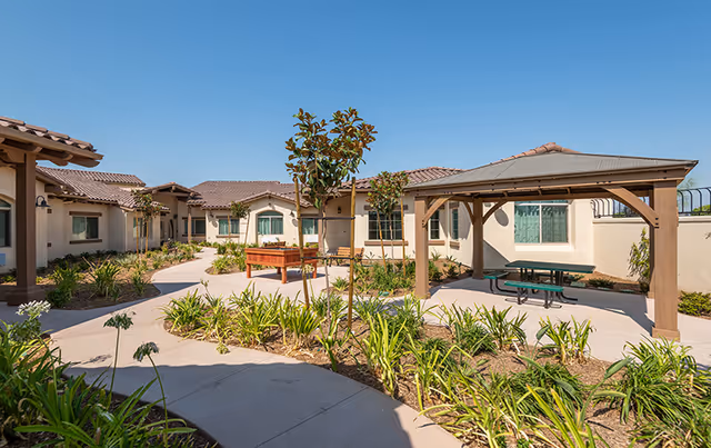 Outdoor courtyard area at ActivCare Orange featuring a paved walkway, landscaped garden beds with plants and small trees, a wooden pergola with a picnic table underneath, and beige stucco buildings with tiled roofs surrounding the space under a clear blue sky.