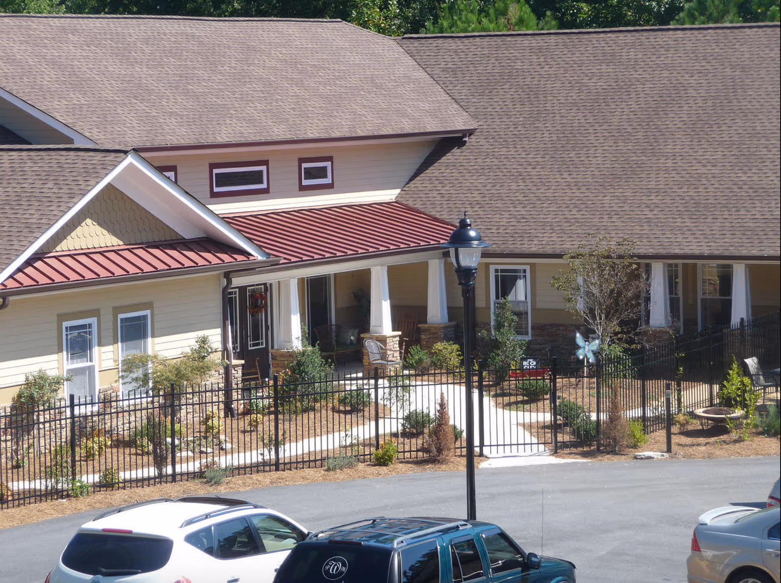 Exterior view of Daybreak Village Assisted Living showing a beige building with a brown shingled roof and a red metal awning over the entrance. There is a black metal fence surrounding a landscaped garden area with shrubs and small trees. Several cars are parked in front of the building on a paved driveway, and a black street lamp is visible near the entrance.