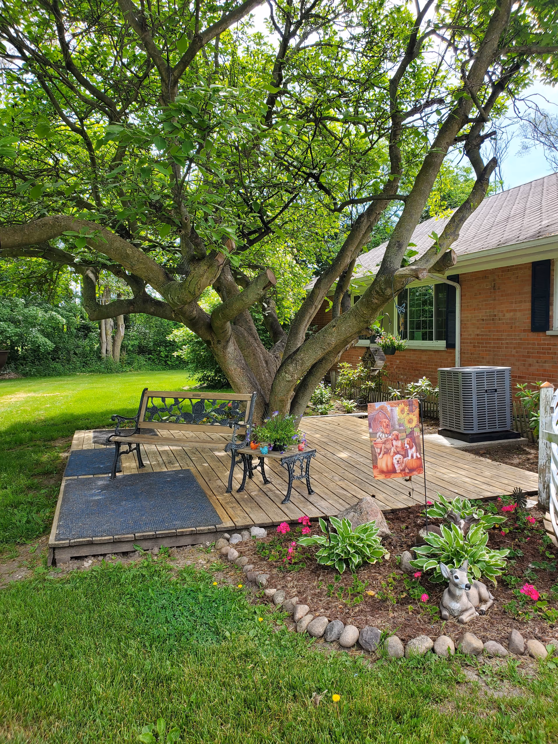 A shaded wooden deck beneath a large tree with a bench and small table beside a brick house, bordered by flowerbeds and lawn.