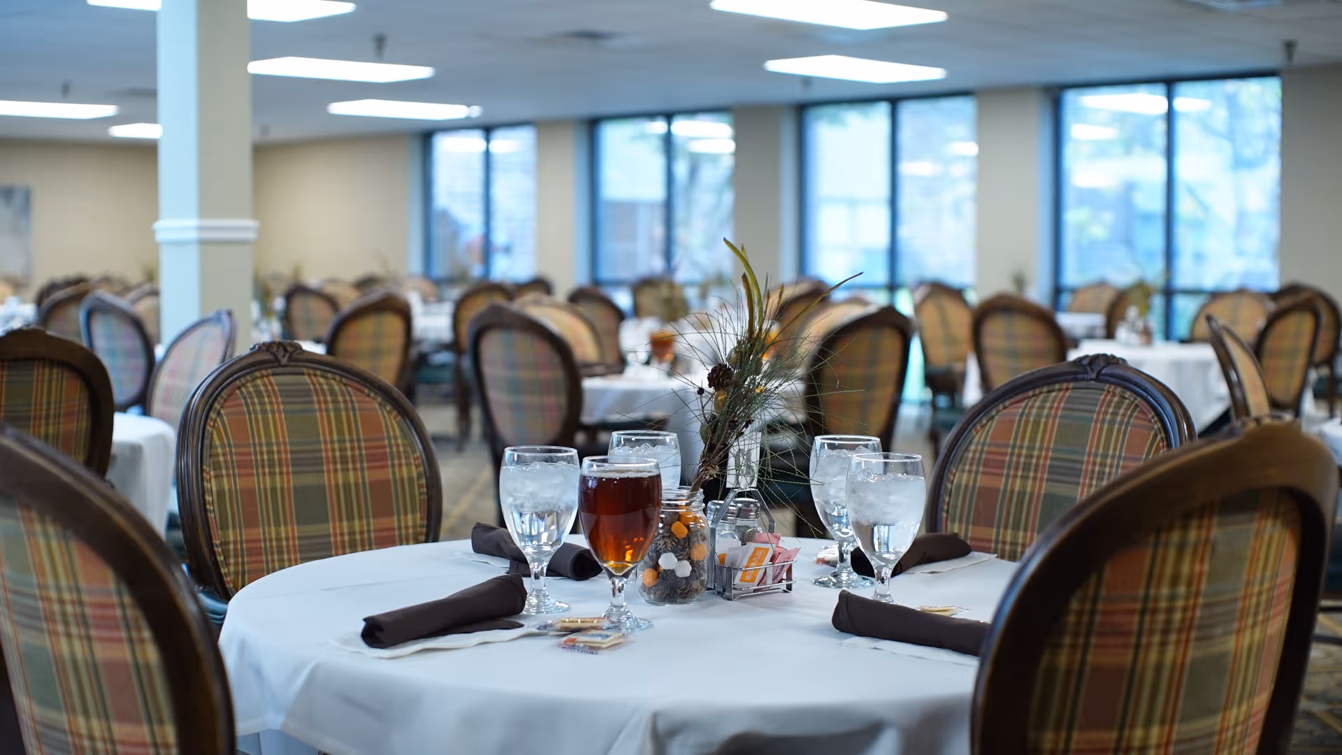 A dining room in a senior living community with round tables covered in white tablecloths. Each table is set with glasses of water and iced tea, dark cloth napkins, and a small centerpiece with decorative foliage. The chairs have wooden frames and plaid upholstery. Large windows in the background let in natural light.
