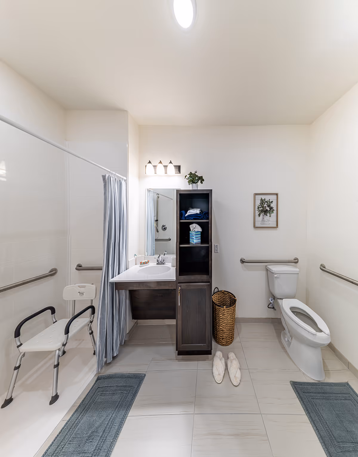 A clean and accessible bathroom with a walk-in shower equipped with a shower chair and grab bars, a vanity with a sink and mirror, a tall dark wood cabinet with towels and tissues, a wicker laundry basket, a toilet with grab bars, a framed floral picture on the wall, and two blue bath mats on the tiled floor. A pair of white slippers is placed on the floor near the cabinet.