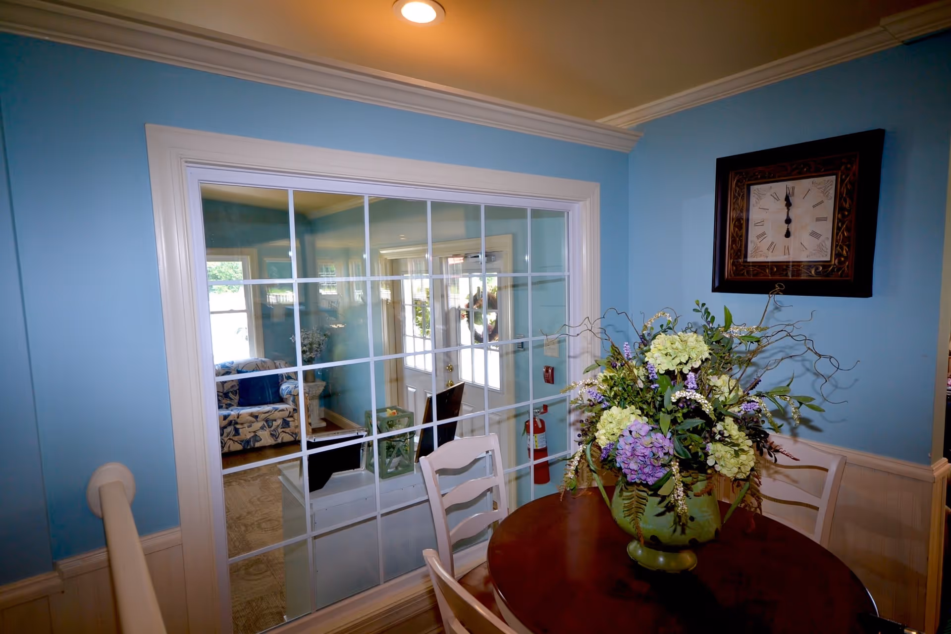 Interior view of a room with light blue walls and white trim, featuring a round wooden table with a large floral arrangement in a green vase. A white chair is partially visible. A large window with white grid framing looks into another room with a patterned armchair and a door with glass panels. A decorative wall clock with Roman numerals hangs on the wall above the table.
