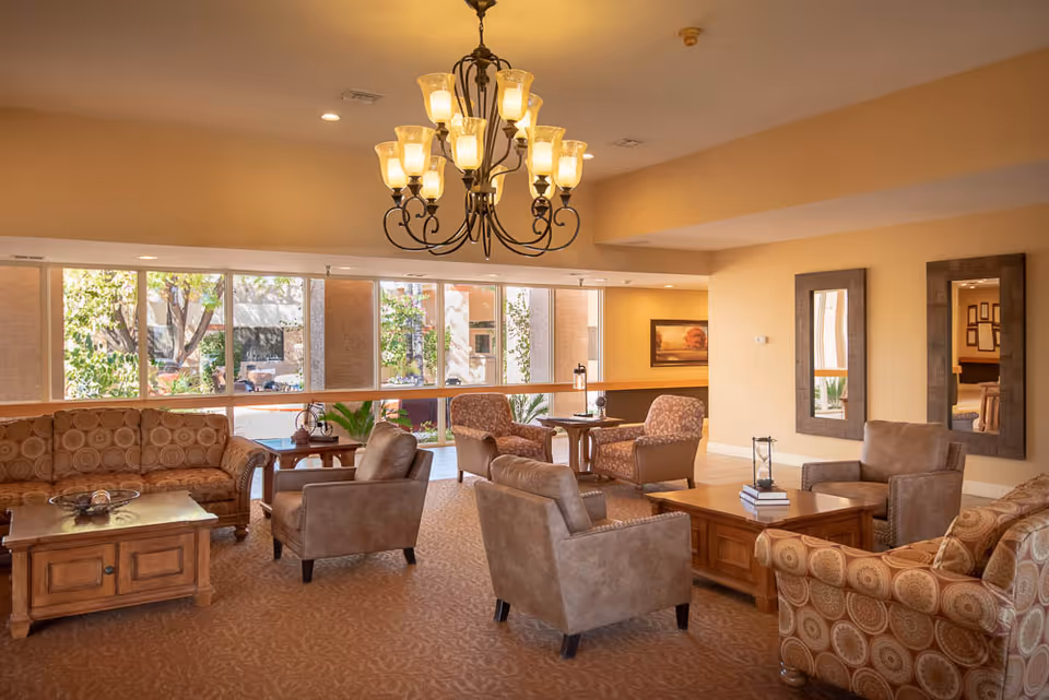 A spacious and well-lit living room area with large windows letting in natural light. The room features multiple comfortable armchairs and patterned sofas arranged around wooden coffee tables. A decorative chandelier hangs from the ceiling, and two large mirrors are mounted on the wall. The room has a warm, inviting atmosphere with beige walls and carpeted flooring.