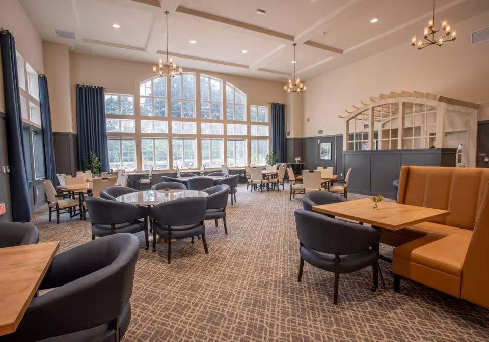 Spacious communal dining and lounge area with round and rectangular tables, upholstered chairs and booths beneath large arched windows and chandeliers.