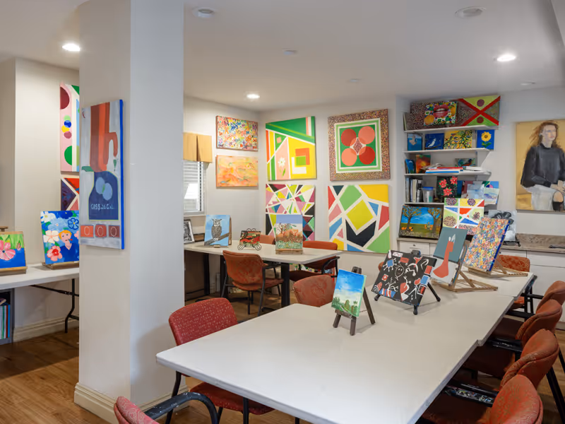 Bright communal art room with tables, chairs, and numerous colorful paintings and canvases displayed on walls and easels.