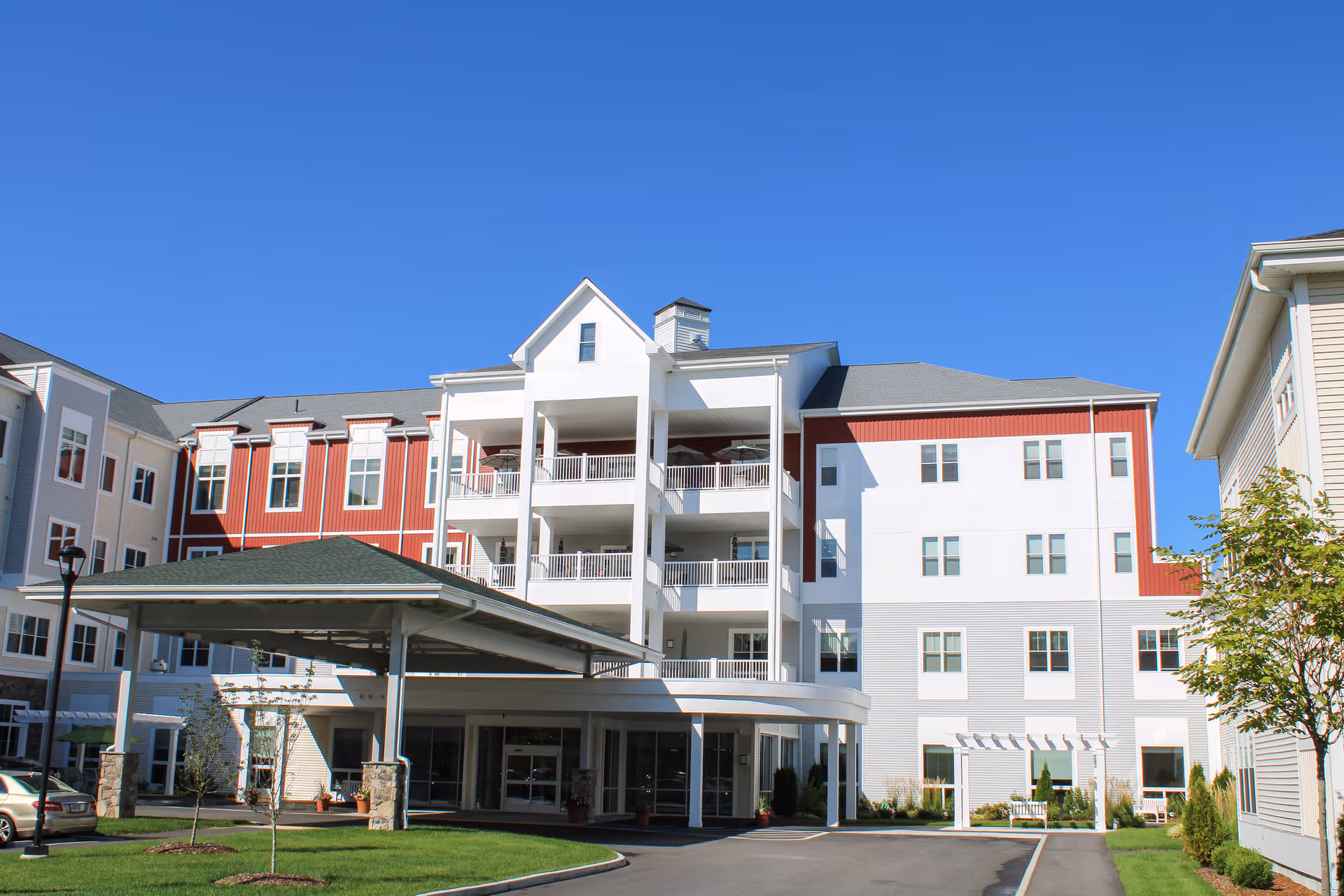 Exterior view of a multi-story assisted living facility building with white and red siding, balconies, a covered entrance, and a clear blue sky.