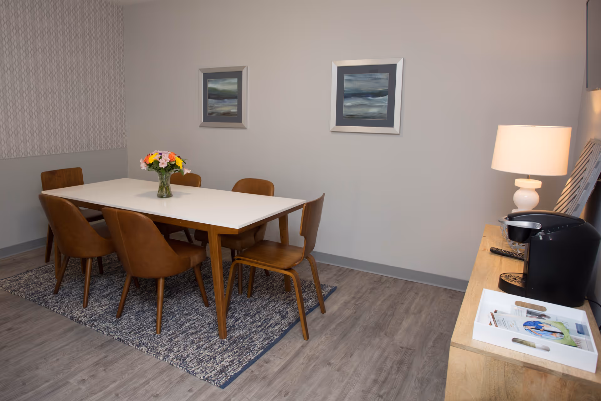 A dining area with a rectangular white table surrounded by six brown chairs on a blue patterned rug. A vase with colorful flowers is placed in the center of the table. On the right side, there is a wooden sideboard with a lamp, a coffee maker, and a white tray holding some papers and items. Two framed abstract paintings hang on the light gray wall behind the table.