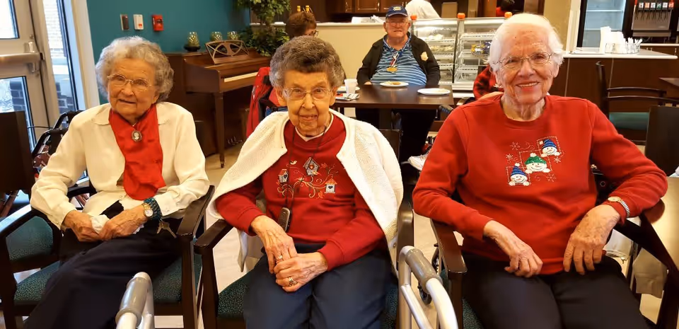 Three elderly women sitting in chairs in a common area of a senior living facility. They are smiling and wearing festive sweaters. Behind them, a man is seated at a table with plates and cups, and there is a piano and a counter with food items in the background.