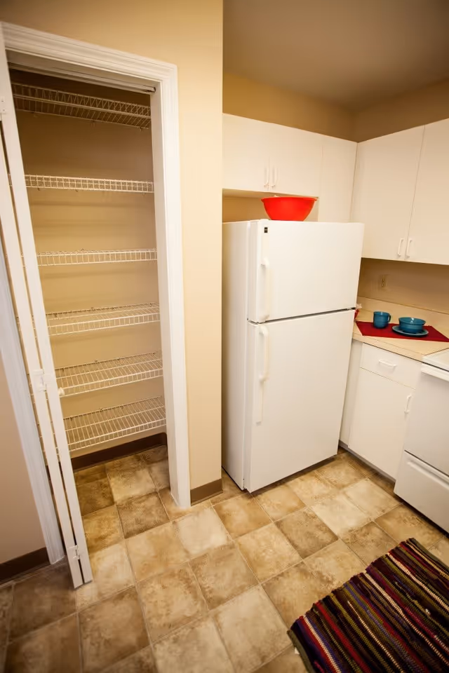 A small kitchen area with a white refrigerator, white cabinets, and a countertop with two blue bowls and plates on a red placemat. To the left is an open pantry with white wire shelves. The floor is covered with beige and brown patterned tiles, and a striped rug is partially visible in the bottom right corner.
