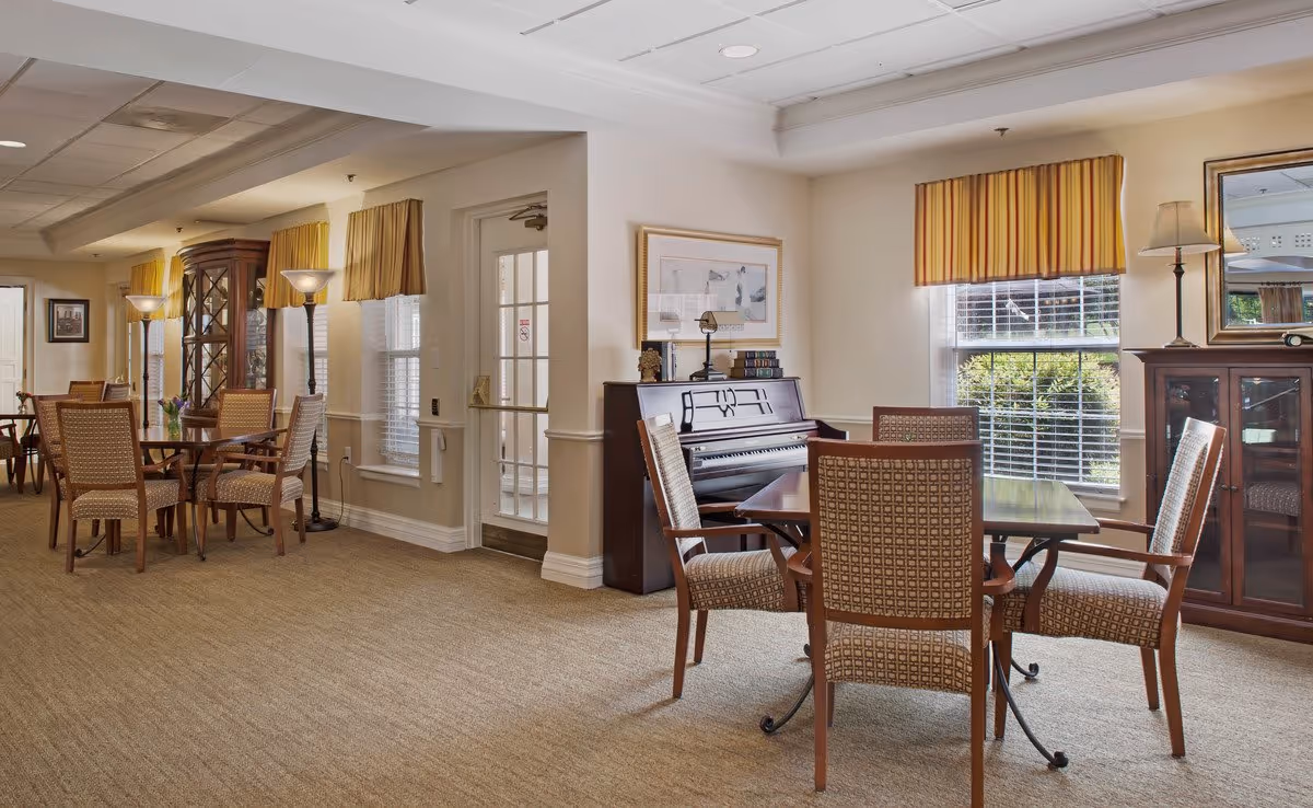Common area with several dining tables and upholstered chairs, a piano and cabinets beneath windows with striped valances.